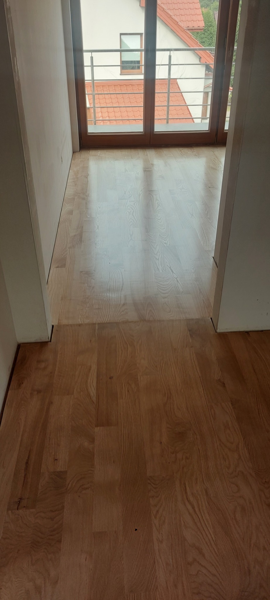 Fragment podłogi z jasnego parkietu drewnianego, przechodzącego w ciemniejszy odcień przy progu, z widocznymi drzwiami balkonowymi w tle.