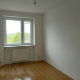 Pusty pokój z drewnianą podłogą, białymi ścianami i oknem z widokiem na zieleń, wisząca żarówka bez klosza u sufitu.