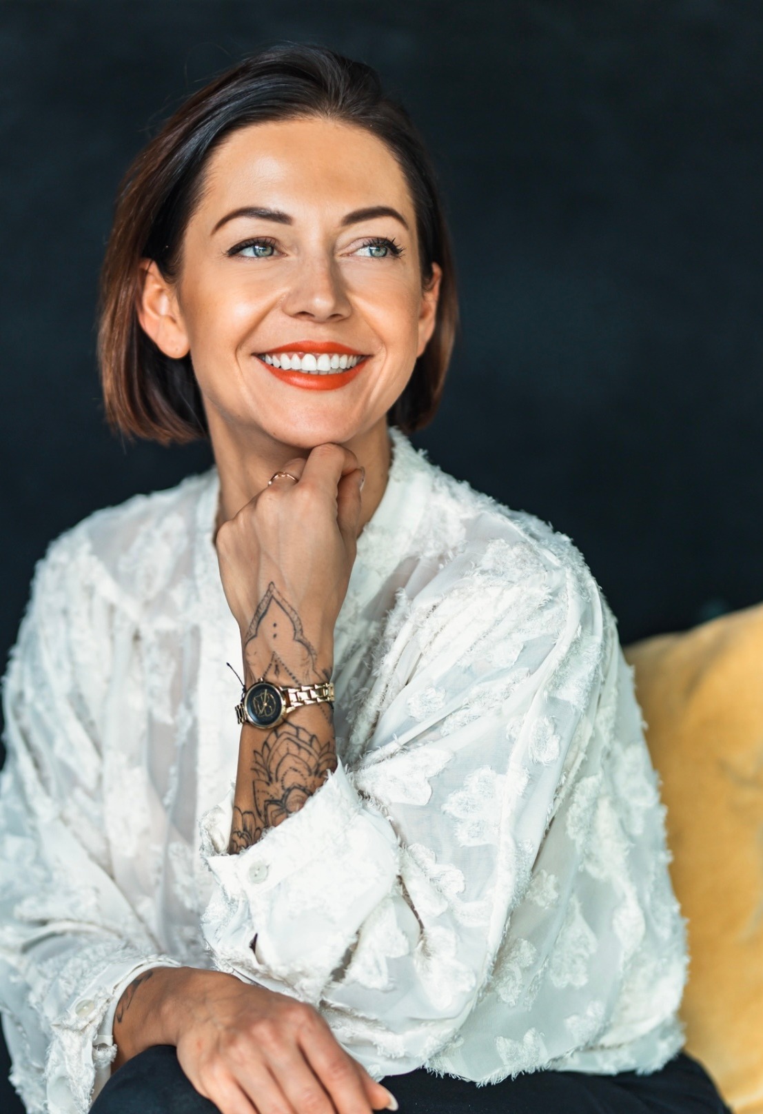 Uśmiechnięta kobieta z krótkimi brązowymi włosami, w białej bluzce o fakturze kwiatowej, z tatuażem na przedramieniu i złotym zegarkiem, opiera podbródek na dłoni z pierścionkiem, spoglądając...