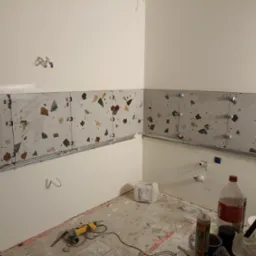 Montaż płytek terazzo na ścianie w trakcie remontu łazienki, widoczne listwy montażowe, krzyżyki dystansowe, puszki elektryczne i narzędzia na podłodze.