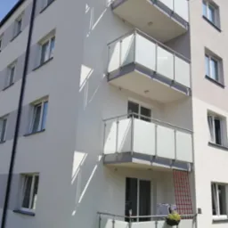 Nowoczesny budynek wielorodzinny z balkonami z mlecznego szkła i metalowymi balustradami, na balkonie na pierwszym piętrze widoczna czerwona siatka zabezpieczająca.