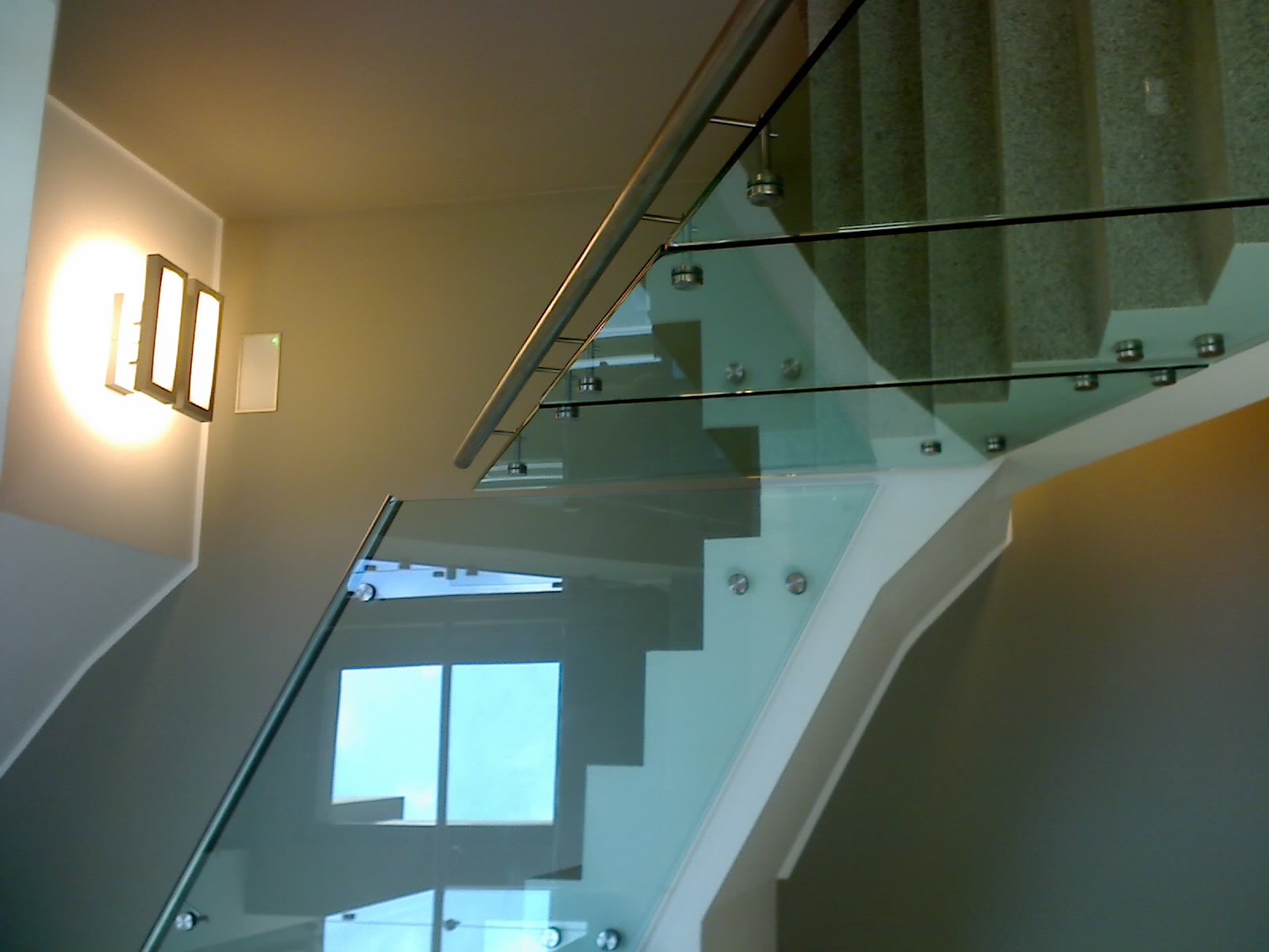 Szklana balustrada z metalowymi elementami mocującymi w Katowicach, widoczna z dołu, z oknem odbijającym światło w tafli szkła.