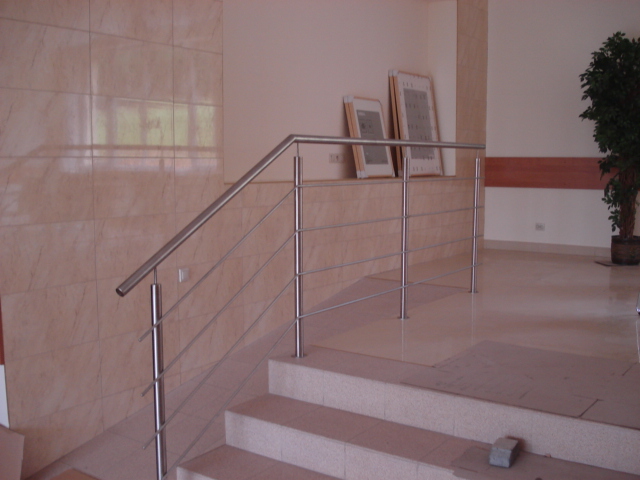 Wewnętrzna balustrada schodowa ze stali nierdzewnej, z prostymi poprzeczkami, na tle jasnych płytek ściennych i podłogowych, z widoczną rośliną doniczkową w tle.