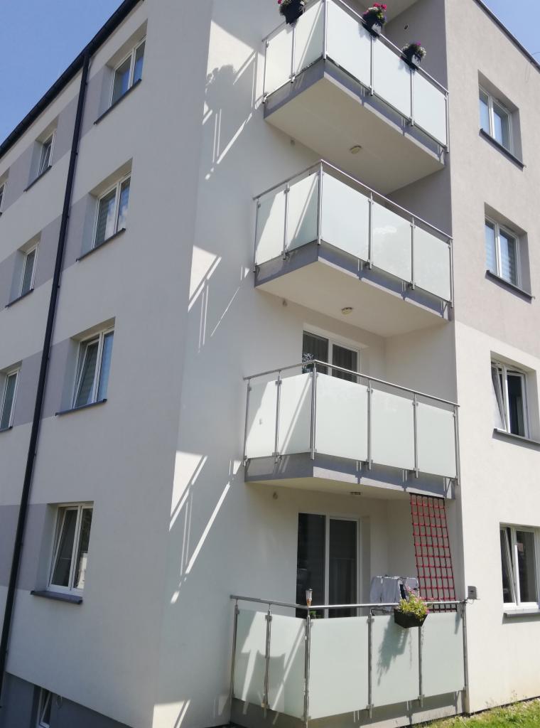 Nowoczesny budynek wielorodzinny z balkonami z mlecznego szkła i metalowymi balustradami, na balkonie na pierwszym piętrze widoczna czerwona siatka zabezpieczająca.