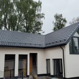 Nowoczesny dom z szarą blachodachówką, czarnymi oknami i jasną elewacją w trakcie wykańczania, widoczne drzewa w tle.
