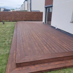 PrimaSort Dach Paweł Walczak - Nowoczesny taras z brązowych desek z dwustopniowymi schodami, otoczony zielonym trawnikiem, z widocznym drewnianym ogrodzeniem i fragmentem elewacji budynku w tle.