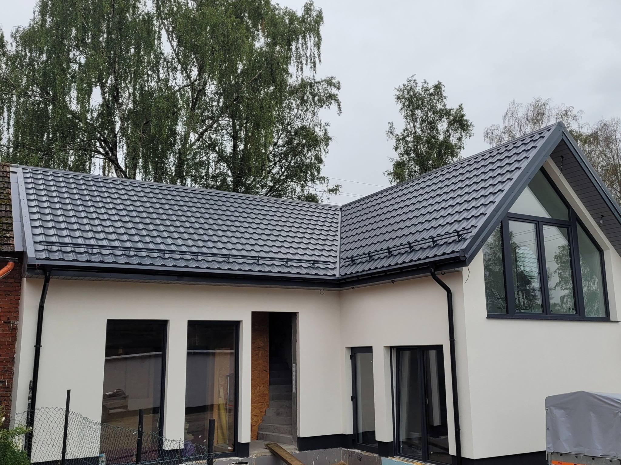 Nowoczesny dom z szarą blachodachówką, czarnymi oknami i jasną elewacją w trakcie wykańczania, widoczne drzewa w tle.