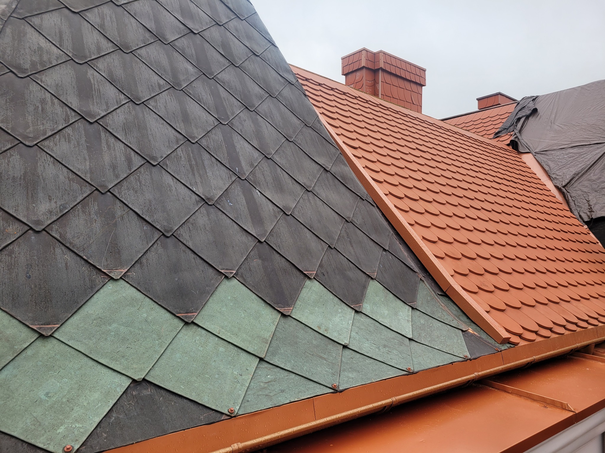 Fragment dachu z łupka w kolorze grafitowym i miedzianym, z widocznym kominem i fragmentem zabezpieczenia folią.