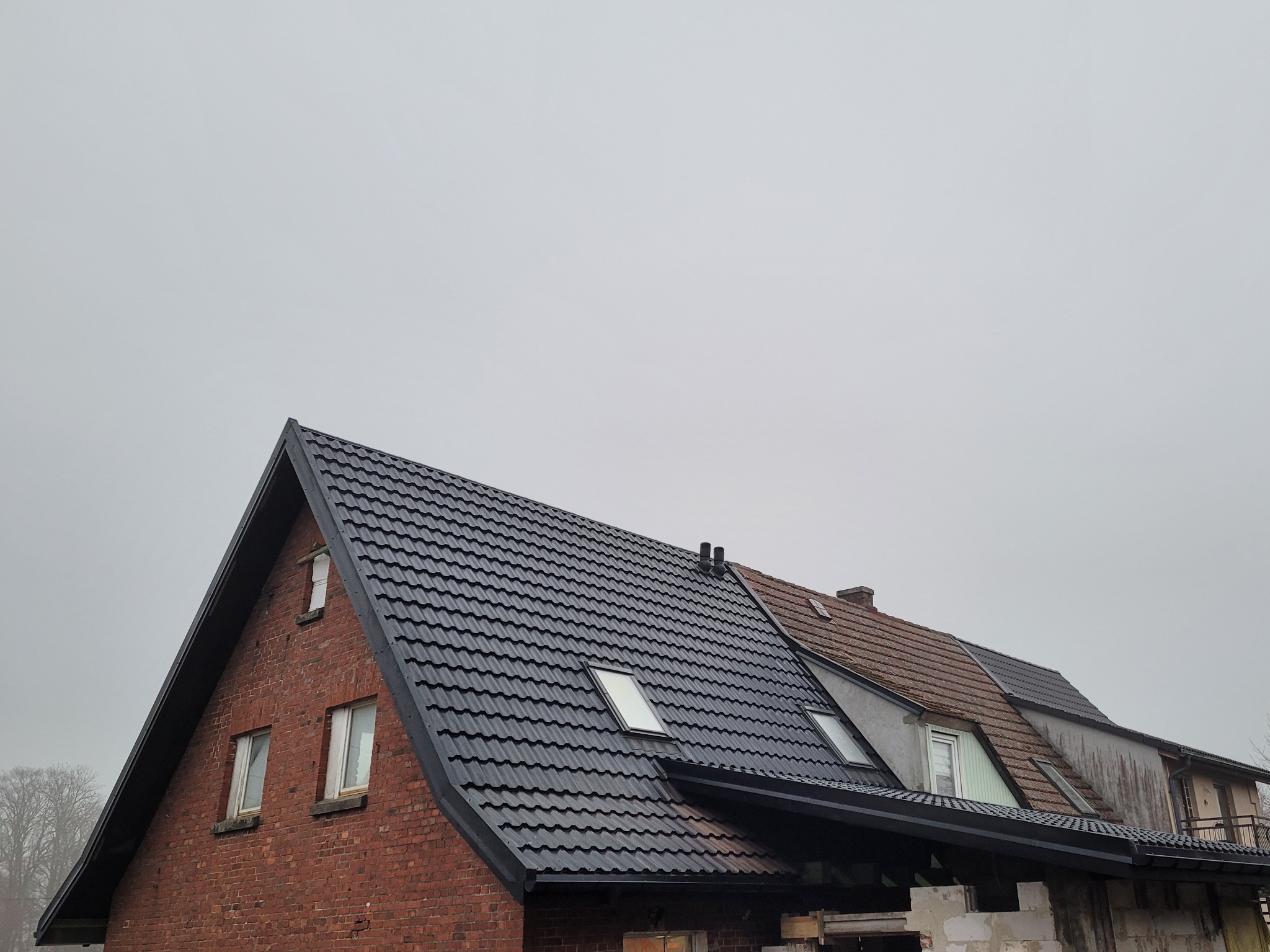 Częściowo wymieniony dach z czarnej blachodachówki na budynku z czerwonej cegły, widoczne okna dachowe i fragment starego dachu z dachówki ceramicznej.