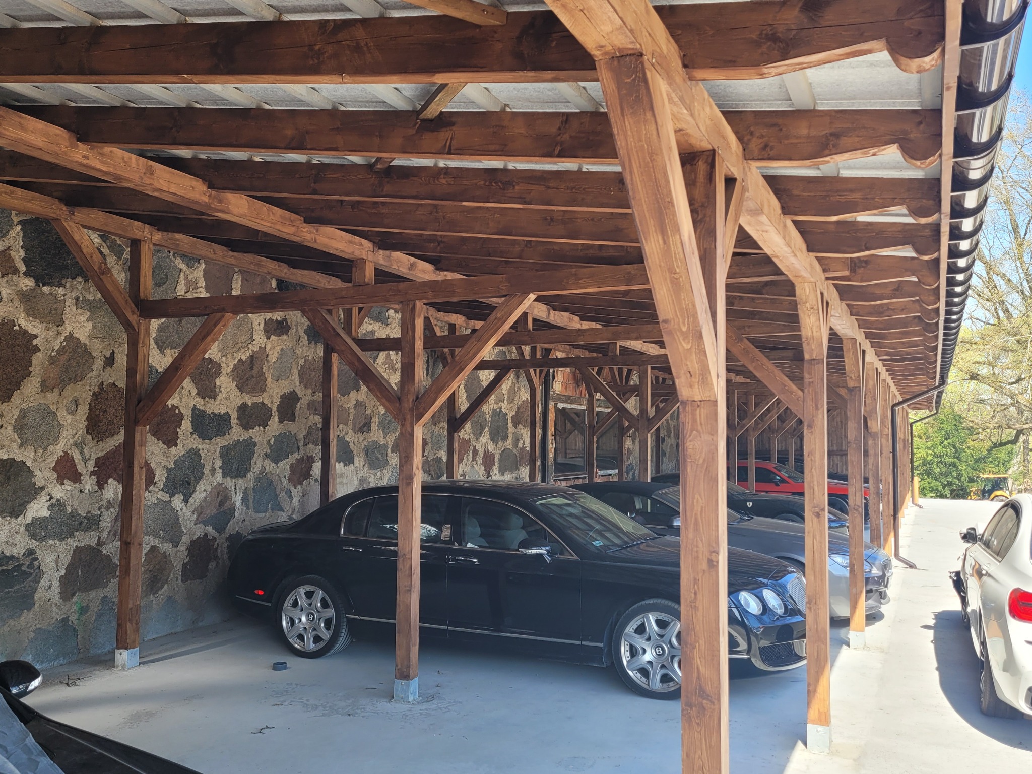 Drewniana wiata garażowa ze ścianami z kamienia polnego w Sławnie, kryta blachą trapezową, z zaparkowanymi samochodami.