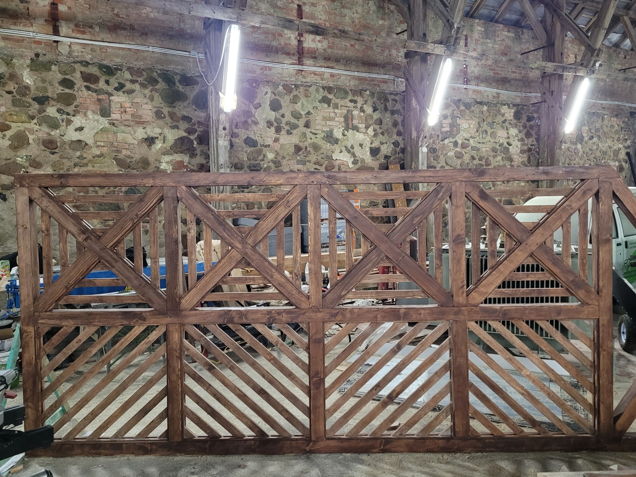Drewniana konstrukcja z kratownicą w kształcie rombów i kwadratów, w odcieniach brązu, na tle kamiennej ściany w pomieszczeniu z widocznymi lampami i elementami warsztatu.