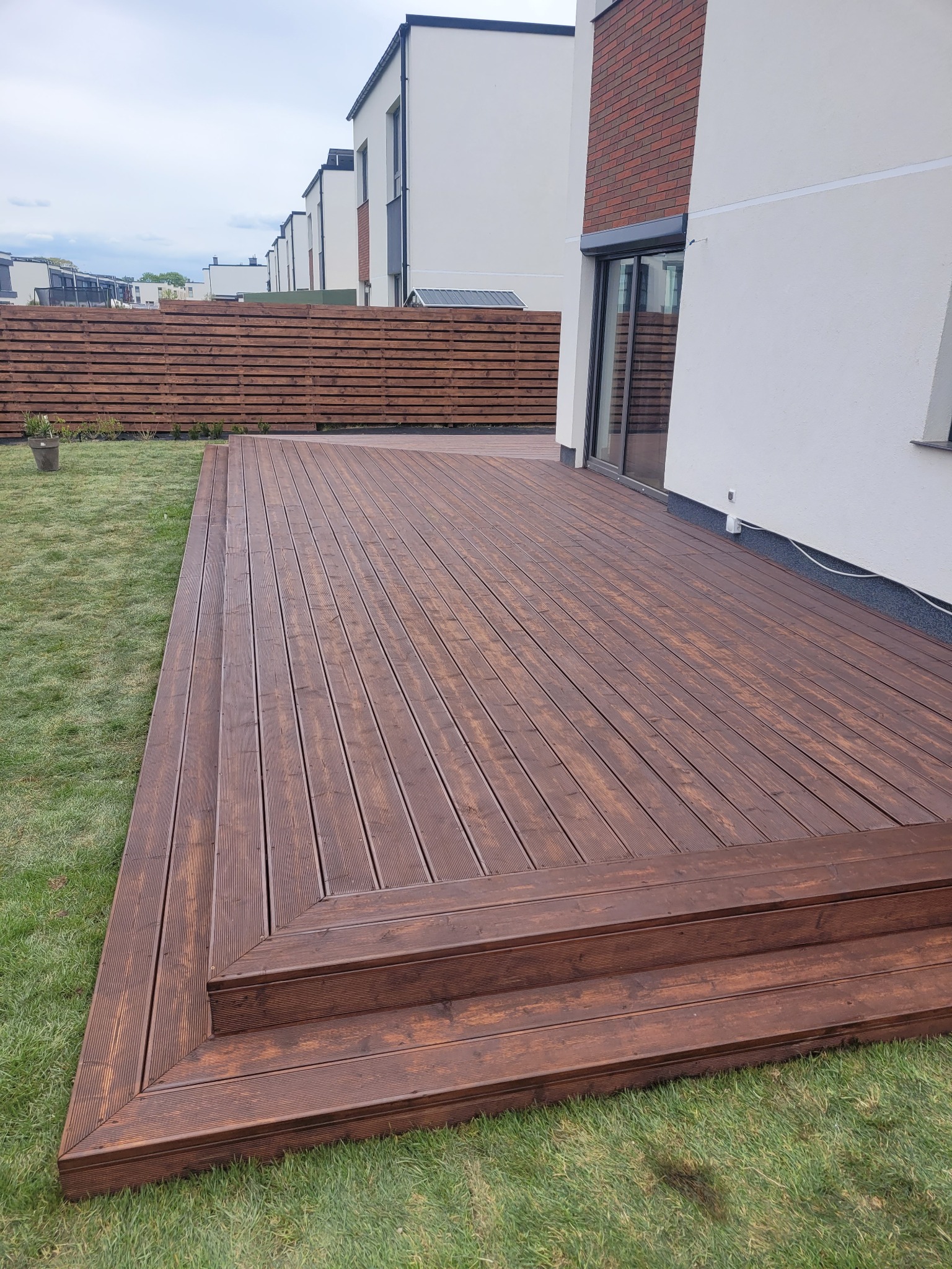 Nowoczesny taras z brązowych desek z dwustopniowymi schodami, otoczony zielonym trawnikiem, z widocznym drewnianym ogrodzeniem i fragmentem elewacji budynku w tle.