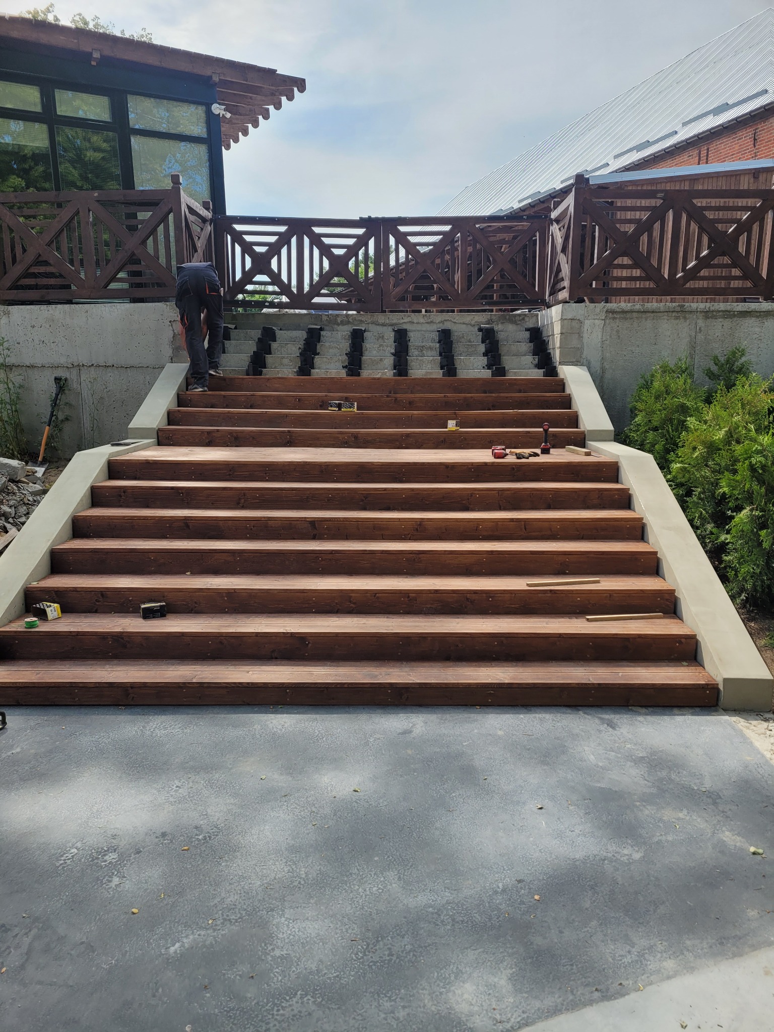 Montaż drewnianych schodów zewnętrznych z betonowymi bokami, widoczne narzędzia i elementy konstrukcyjne, w tle budynek z drewnianą balustradą.