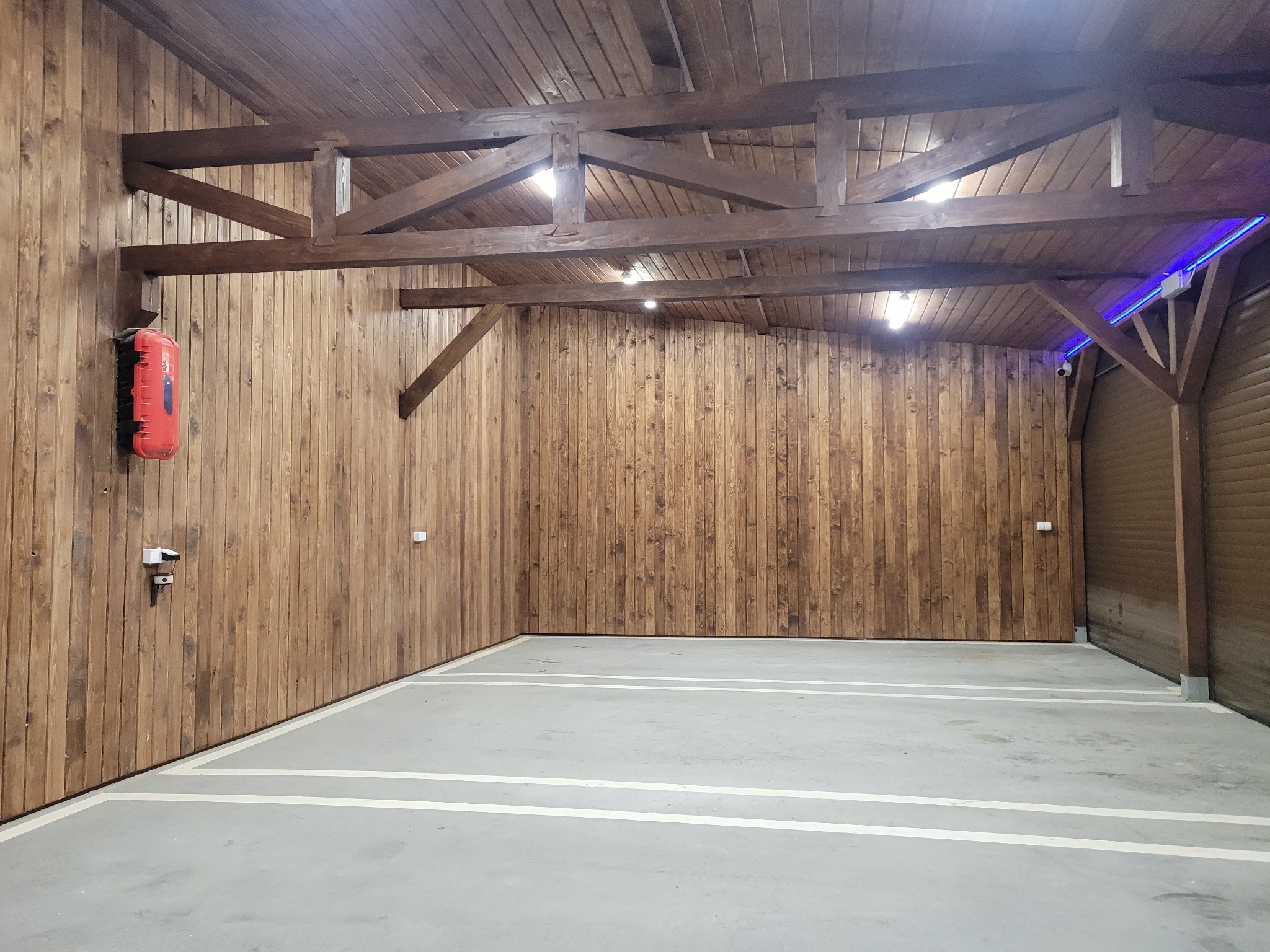 Wnętrze garażu z drewnianą boazerią na ścianach i suficie, widoczne drewniane belki konstrukcyjne, betonowa podłoga z namalowanymi liniami parkingowymi, czerwona gaśnica na ścianie.
