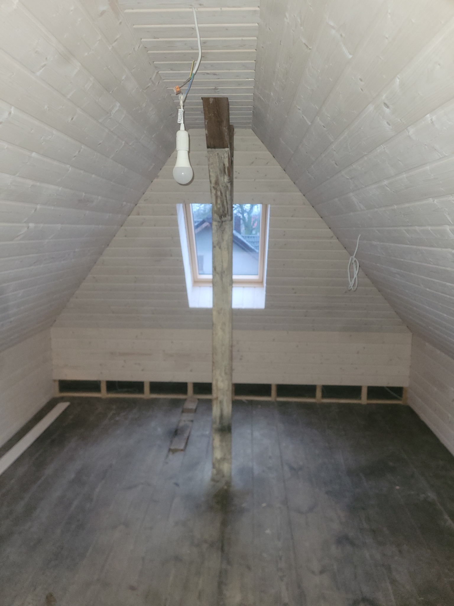 Poddasze z oknem dachowym, wykończone jasnym drewnem boazeryjnym, z widoczną konstrukcją nośną i prowizorycznym oświetleniem w postaci żarówki na kablu.