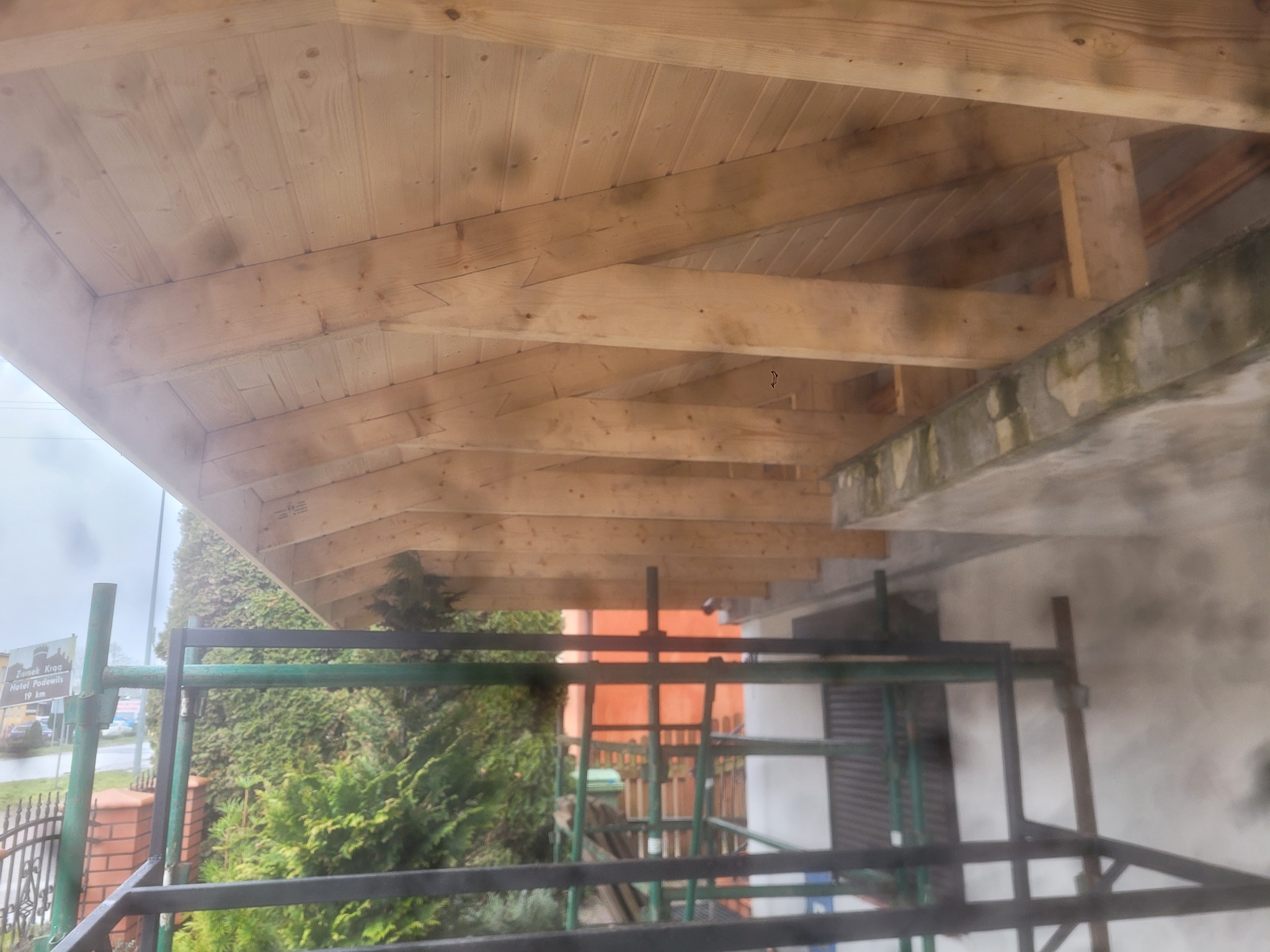 Drewniana konstrukcja dachu od wewnątrz, widoczne deski i krokwie, rusztowanie na pierwszym planie, fragment elewacji budynku w tle.