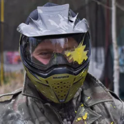 Uczestnik paintballu w pełnym ekwipunku ochronnym, z żółtą farbą na wizjerze maski, ubrany w kurtkę moro.