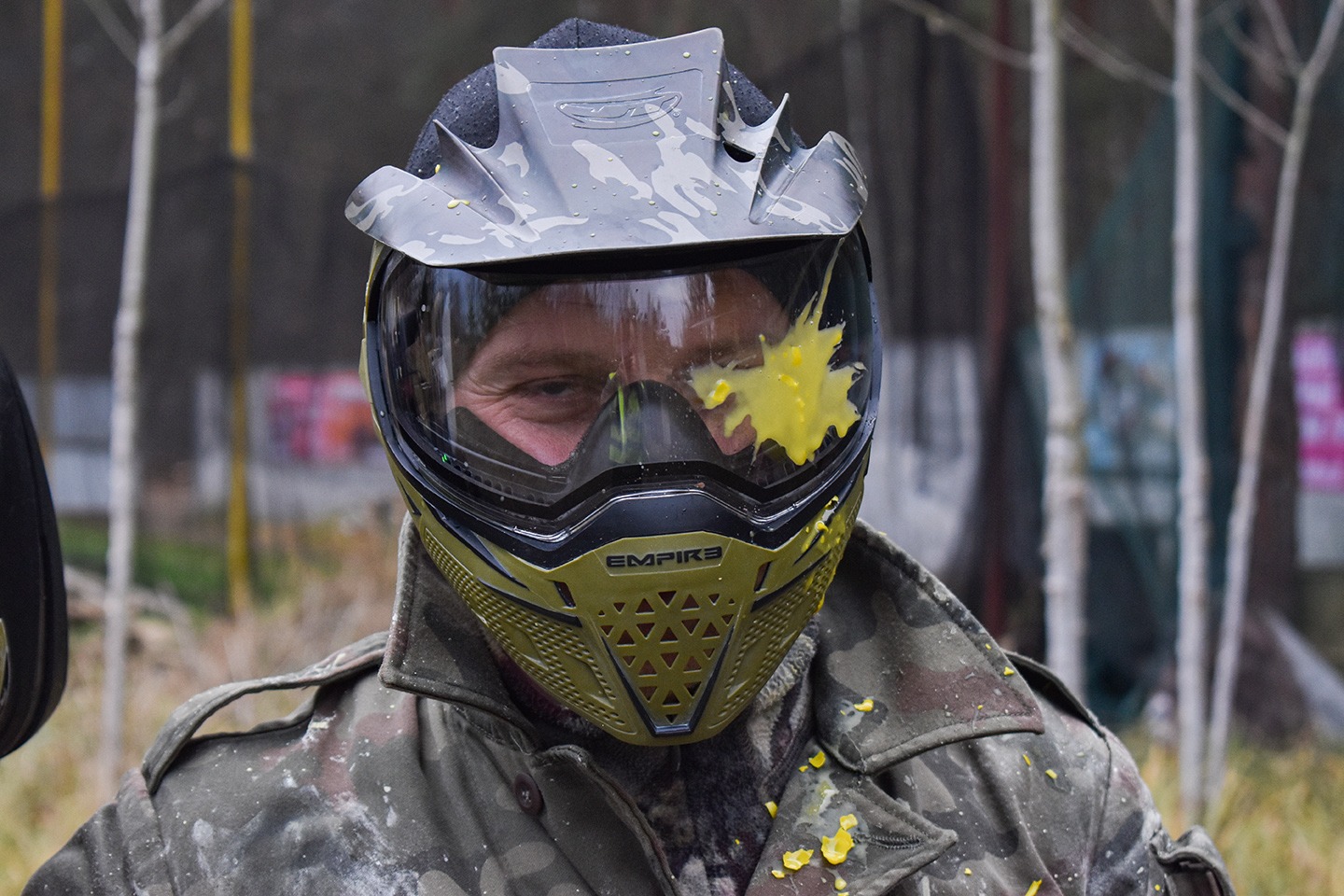 Uczestnik paintballu w pełnym ekwipunku ochronnym, z żółtą farbą na wizjerze maski, ubrany w kurtkę moro.