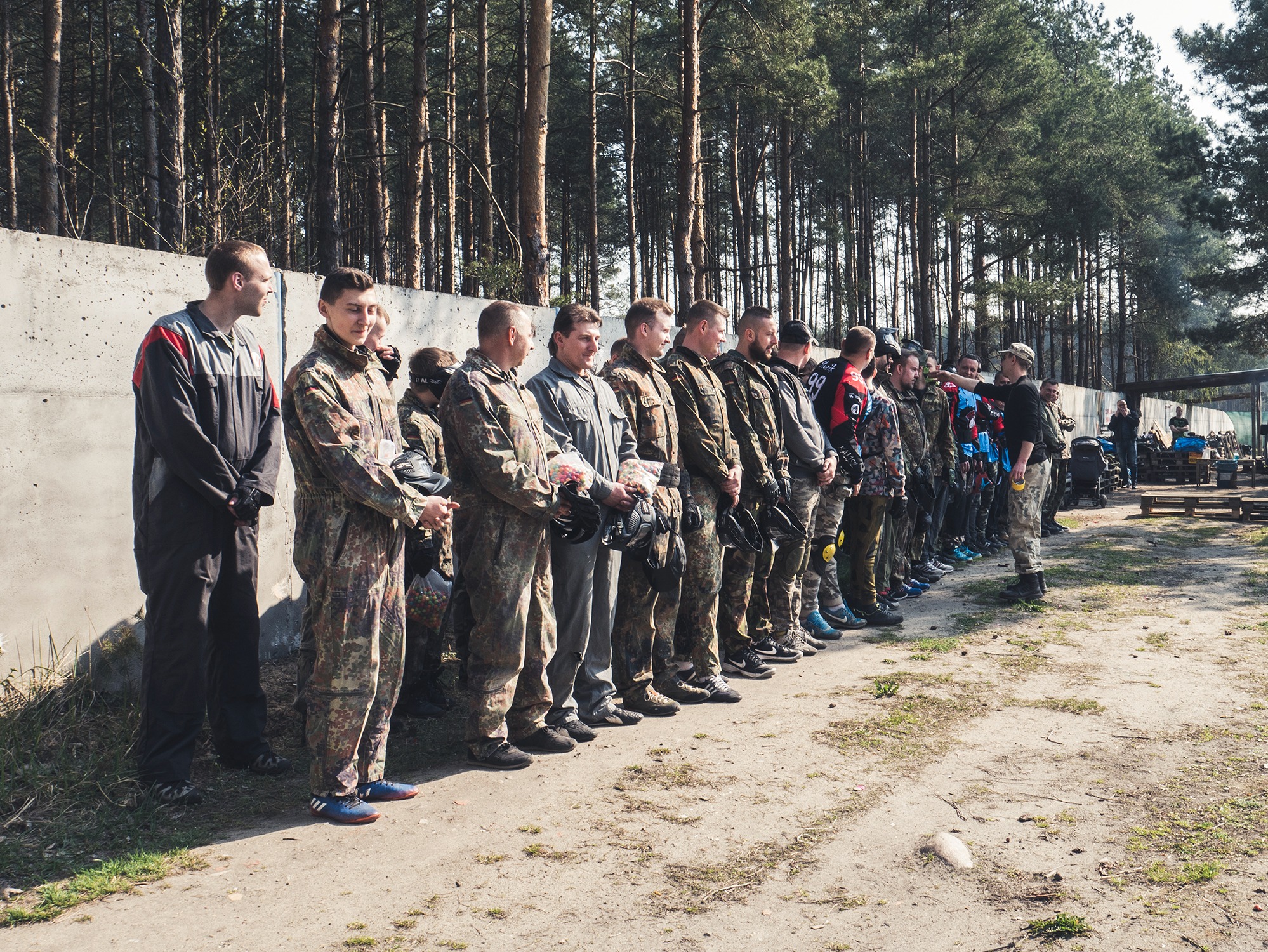 Grupa osób w strojach moro ustawiona w rzędzie, prawdopodobnie przed rozgrywką paintballową, z lasem w tle i instruktorem wskazującym na coś.