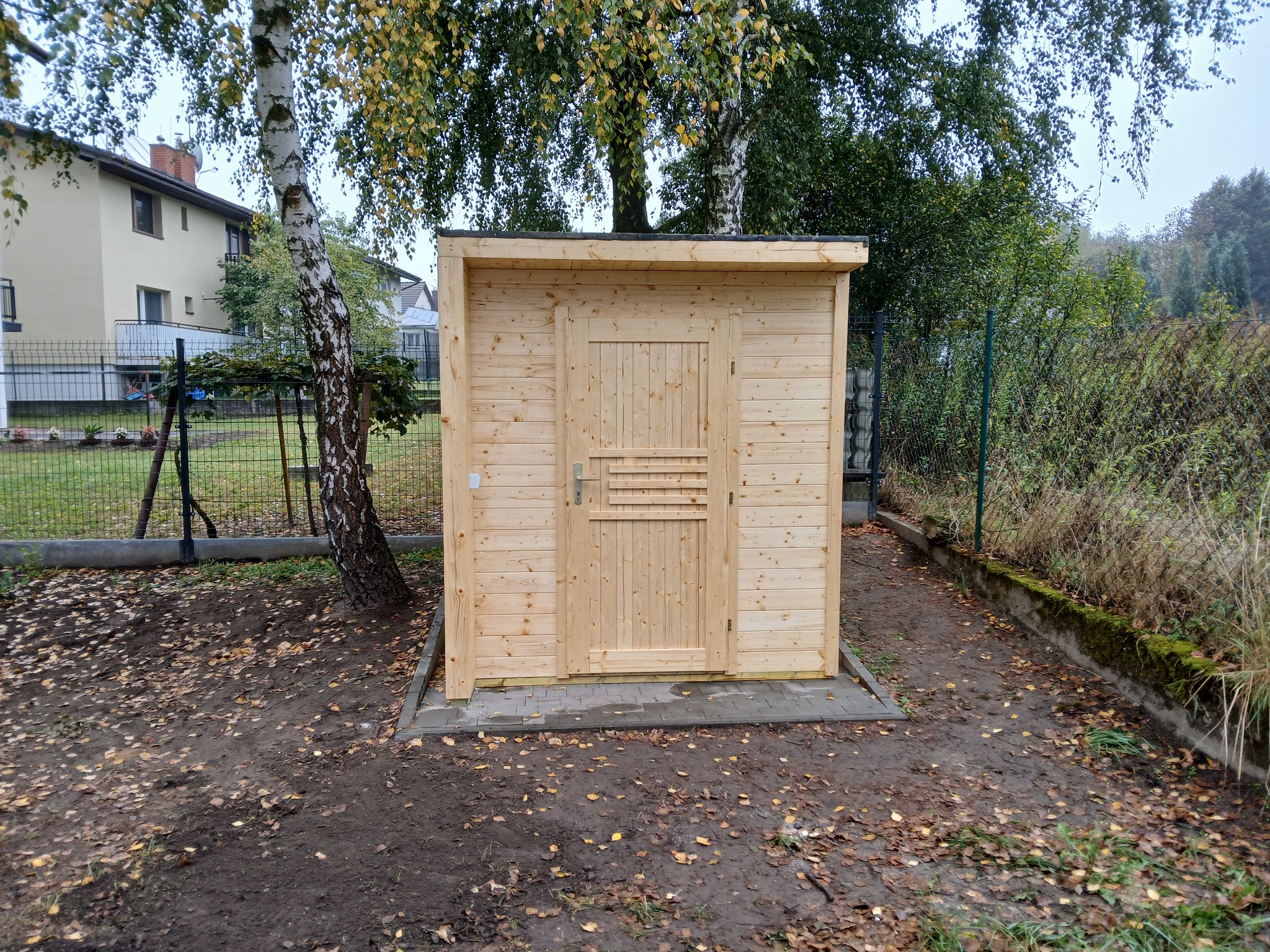 Nowy, drewniany domek narzędziowy z prostą, jednospadową konstrukcją, umieszczony na kostce brukowej, otoczony drzewami i opadłymi liśćmi.