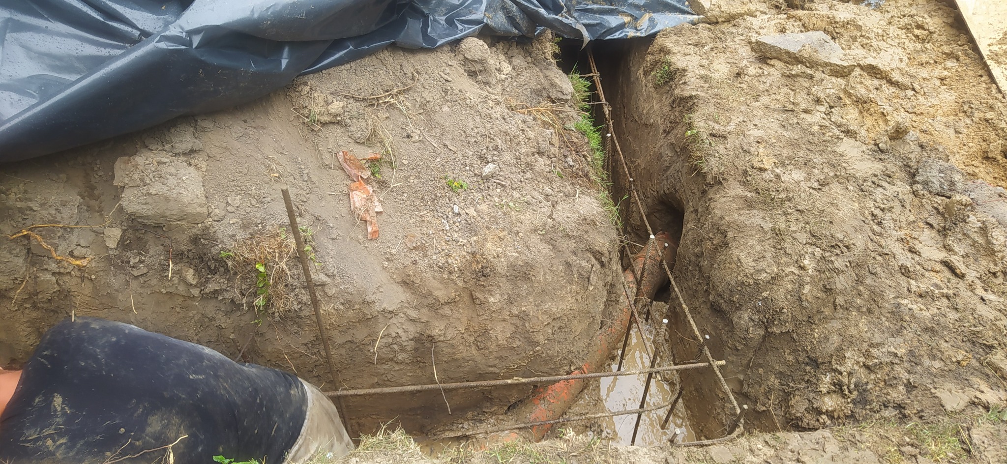 Wykop z widoczną rurą w ziemi, zabezpieczony folią i elementami zbrojeniowymi, przygotowany do dalszych prac ziemnych.