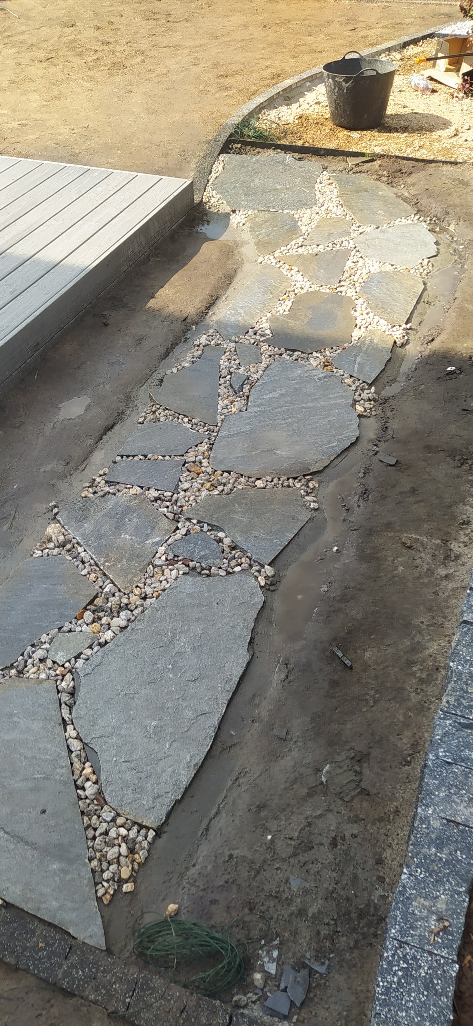 Nieregularne płyty kamienne ułożone jako ścieżka, wypełnione drobnymi kamykami, na wilgotnej, ubitej ziemi, obok drewnianego tarasu.
