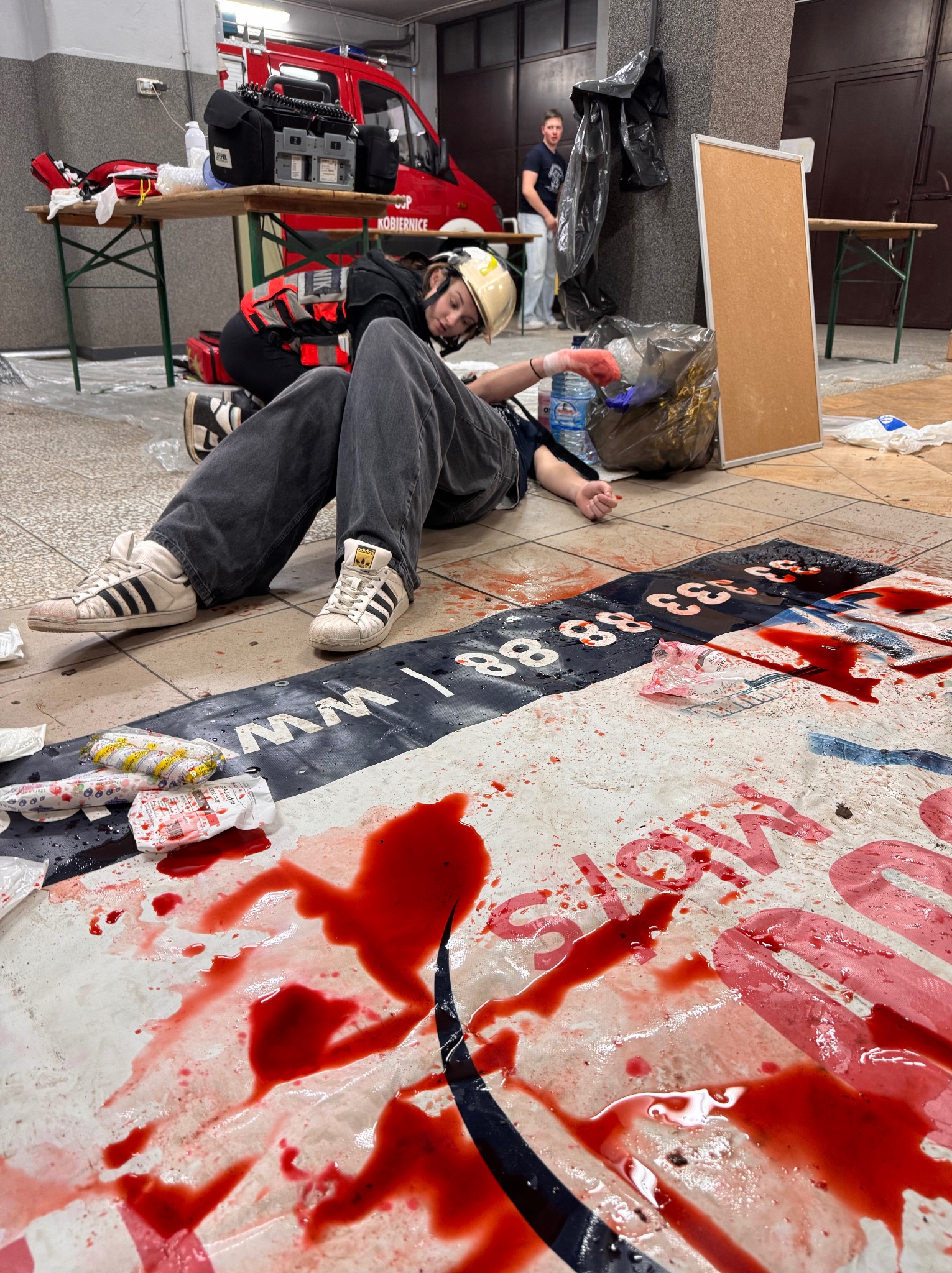 Symulacja udzielania pierwszej pomocy z użyciem sztucznej krwi na macie edukacyjnej. Osoba w kasku i kamizelce udziela pomocy poszkodowanemu. W tle ambulans i wyposażenie.