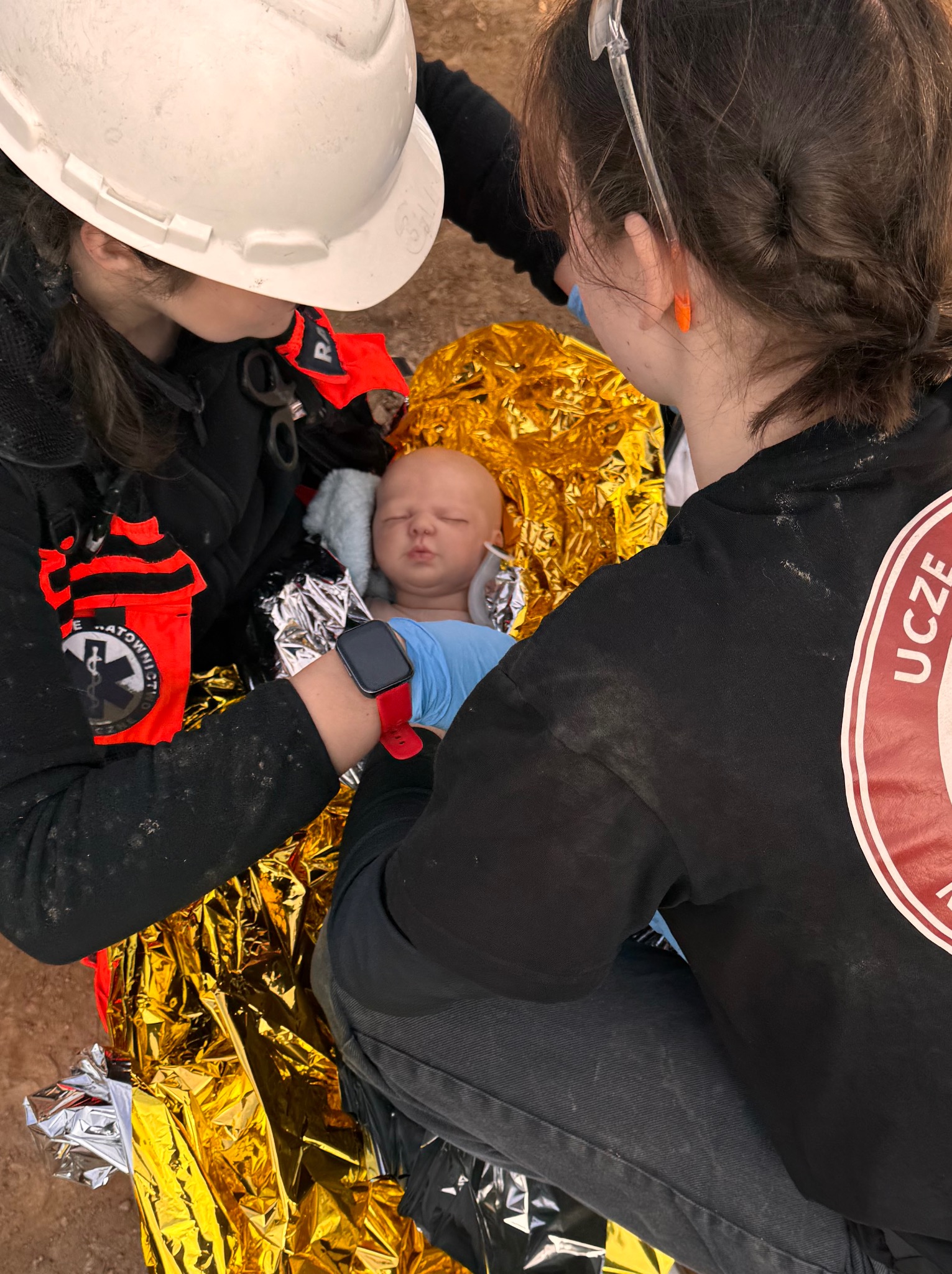 Kurs pierwszej pomocy: Instruktorzy z hełmem i odblaskami, uczą udzielania pierwszej pomocy na manekinie niemowlęcia owiniętym w koc termiczny.