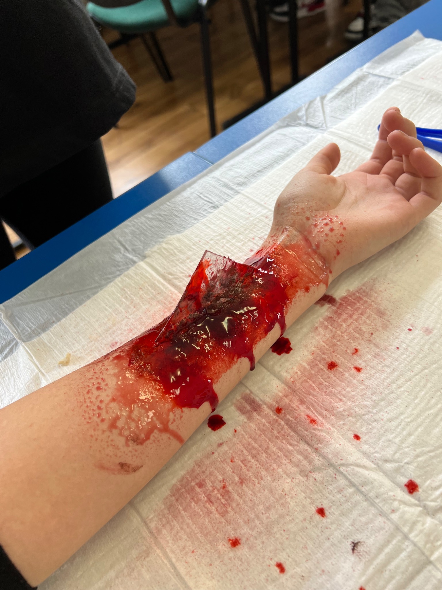 Realistyczna symulacja rany ciętej na przedramieniu, używana podczas zajęć praktycznych z pierwszej pomocy. Krew i uszkodzona skóra na białym podkładzie.