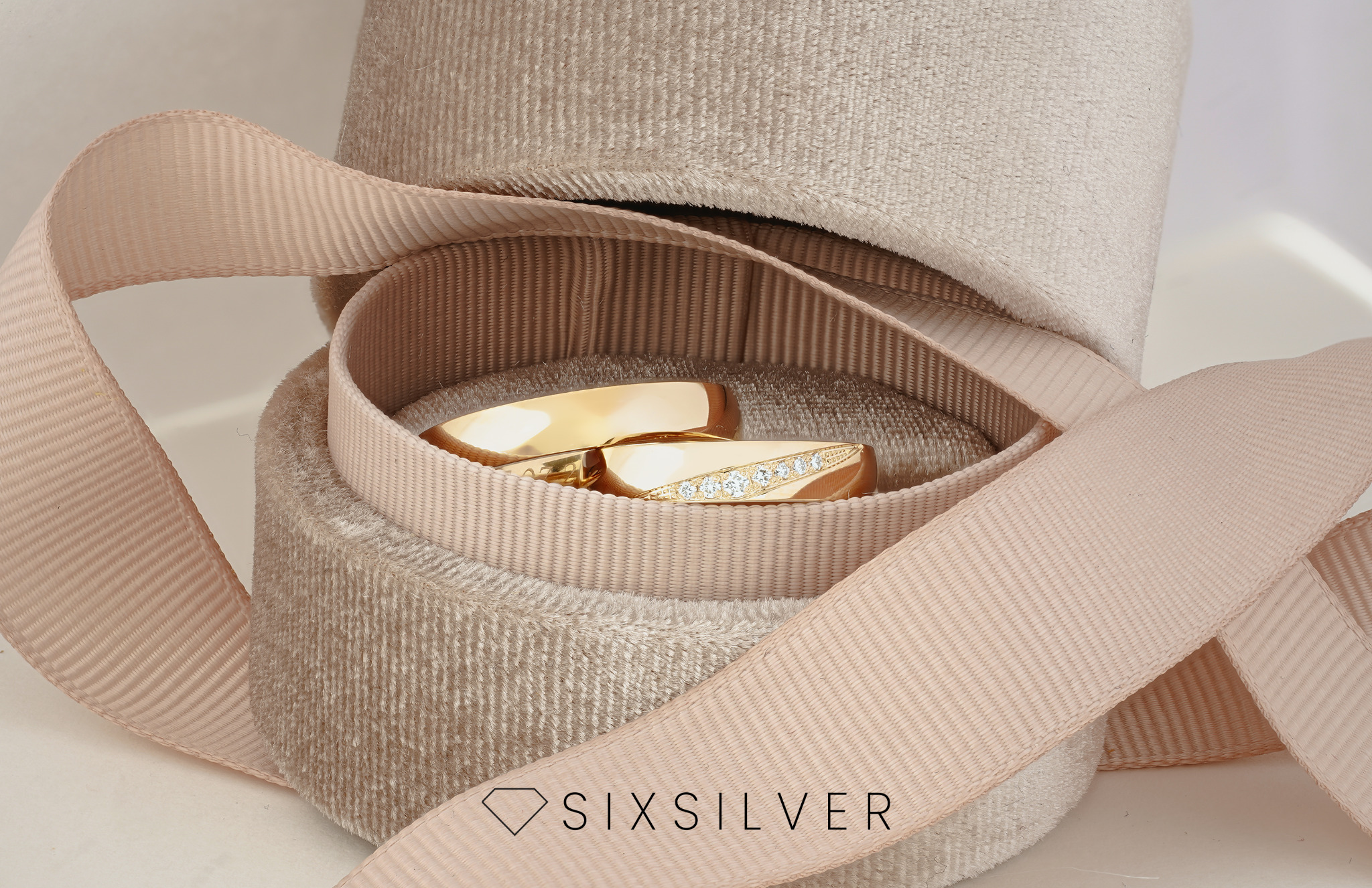 Dwa złote pierścionki, jeden z rzędem małych diamentów, leżące w okrągłym, beżowym pudełku wyściełanym aksamitem, owiniętym satynową wstążką w kolorze pudrowego różu. W dolnej części widoczne logo...