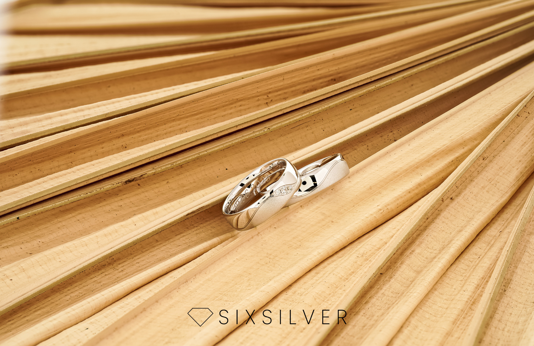 Dwa srebrne pierścionki, jeden z małymi diamentami, leżą na tle suszonych liści palmowych, widoczne logo SixSilver.
