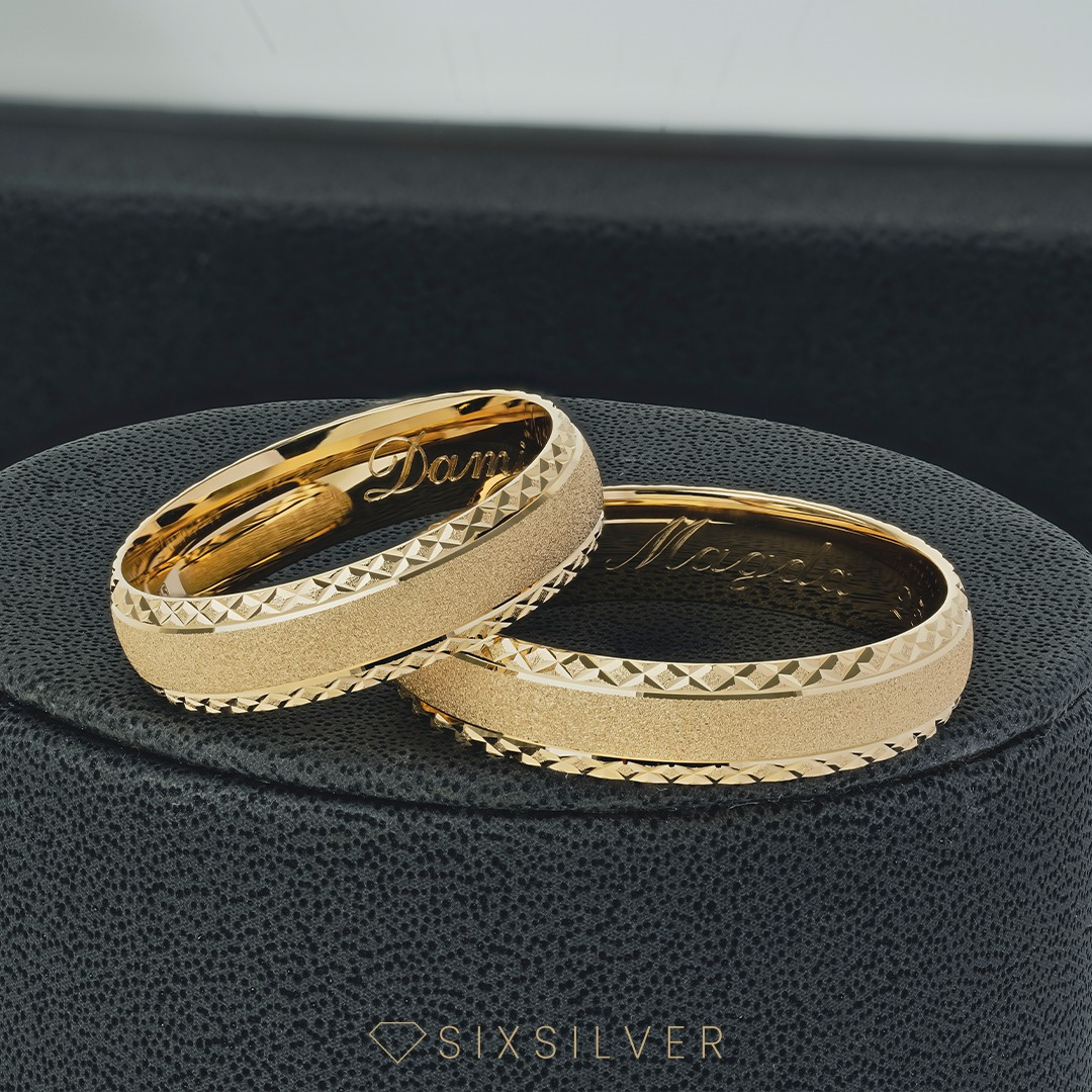 Dwa złote, grawerowane obrączki z imionami 'Damian' i 'Magda' na czarnym, teksturowanym tle, delikatne światło podkreśla detale.
