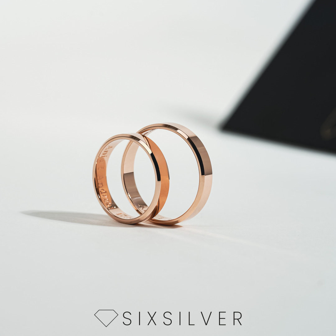 Dwa różowe złote pierścionki z wygrawerowanymi napisami na wewnętrznej stronie, ustawione na białym tle z czarnym pudełkiem w tle, logo SIXSILVER na dole.