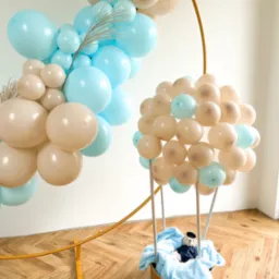 Dekoracja balonowa w kształcie balonu na gorące powietrze z misiem w koszu, obok łuk z balonów w kolorach beżowym i błękitnym, na podłodze parkiet w jodełkę.