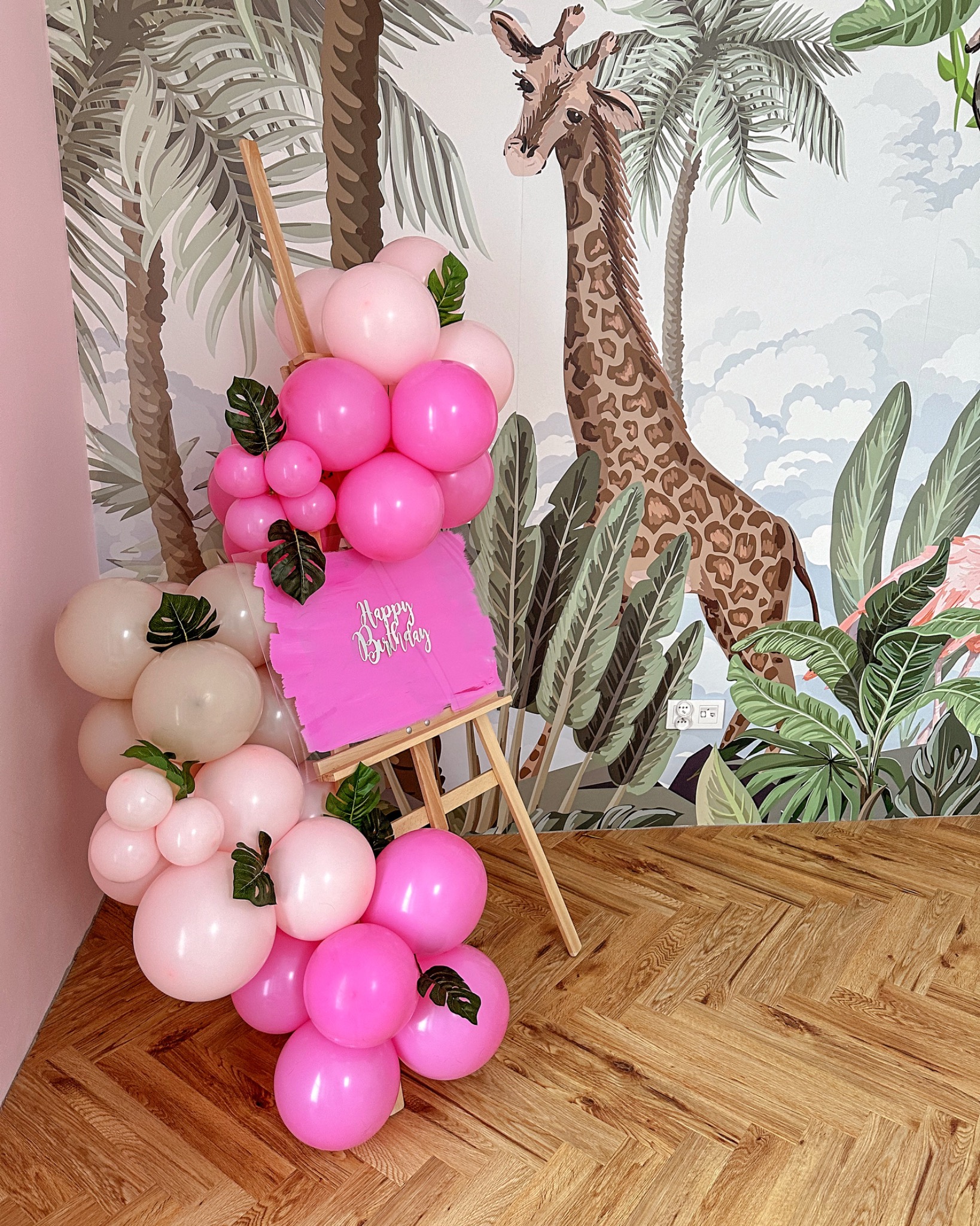 Dekoracja urodzinowa z różowych i białych balonów, drewniany stojak z napisem 'Happy Birthday' i motywem tropikalnym na ścianie z żyrafą.
