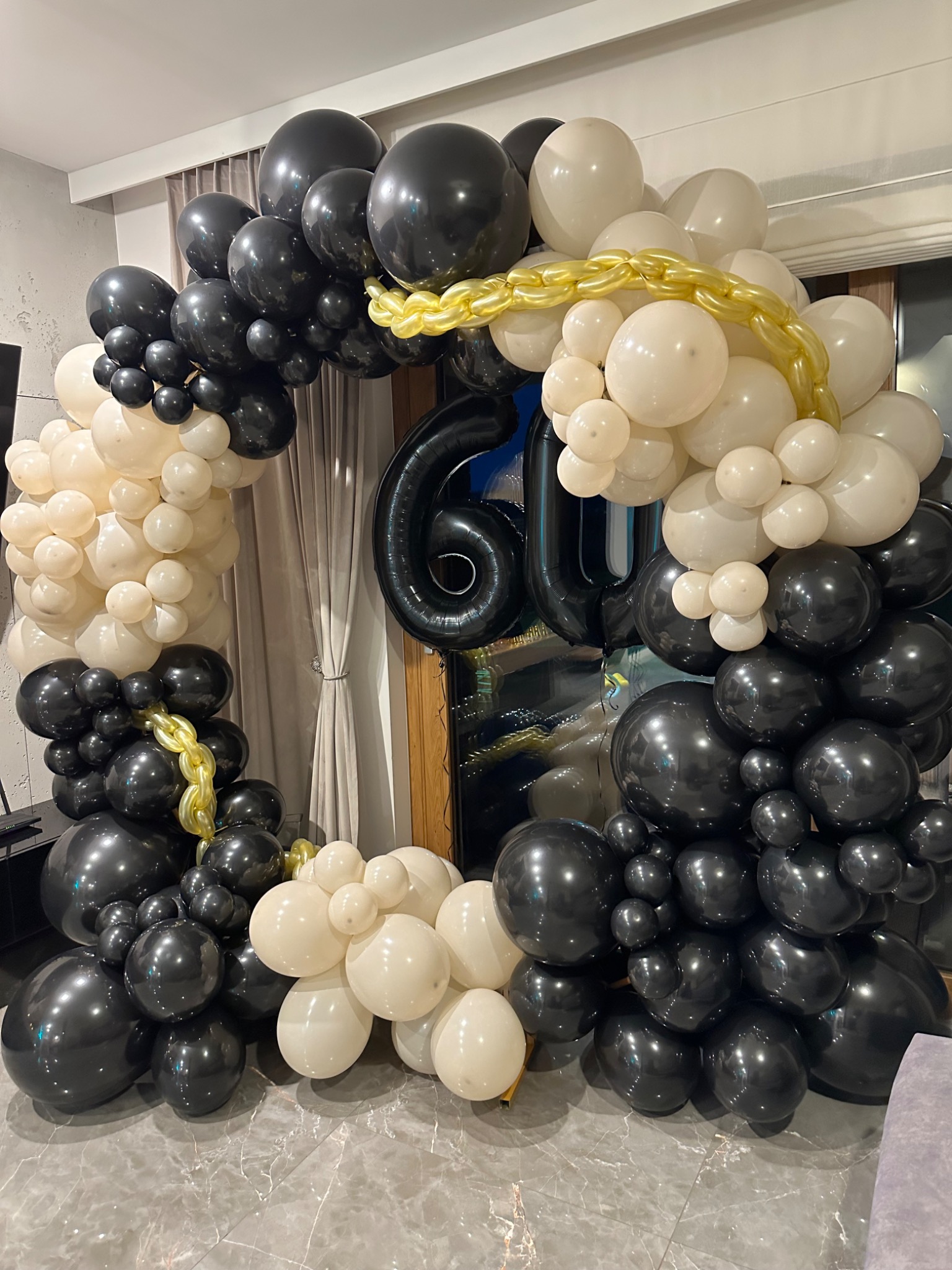 Czarno-biała dekoracja balonowa w kształcie łuku z cyfrą 60 w tle, połączona złotym łańcuchem z balonów.
