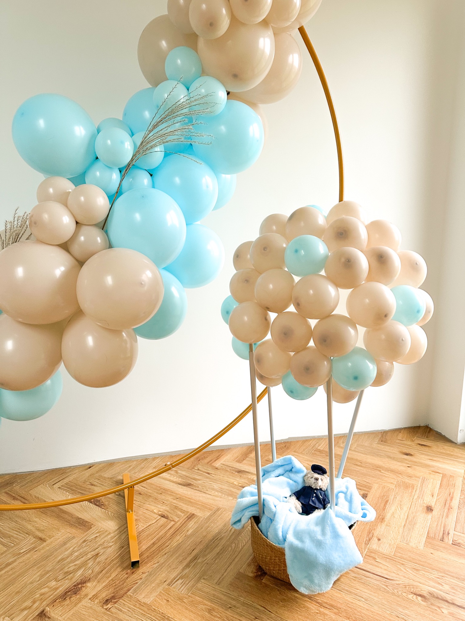 Dekoracja balonowa w kształcie balonu na gorące powietrze z misiem w koszu, obok łuk z balonów w kolorach beżowym i błękitnym, na podłodze parkiet w jodełkę.