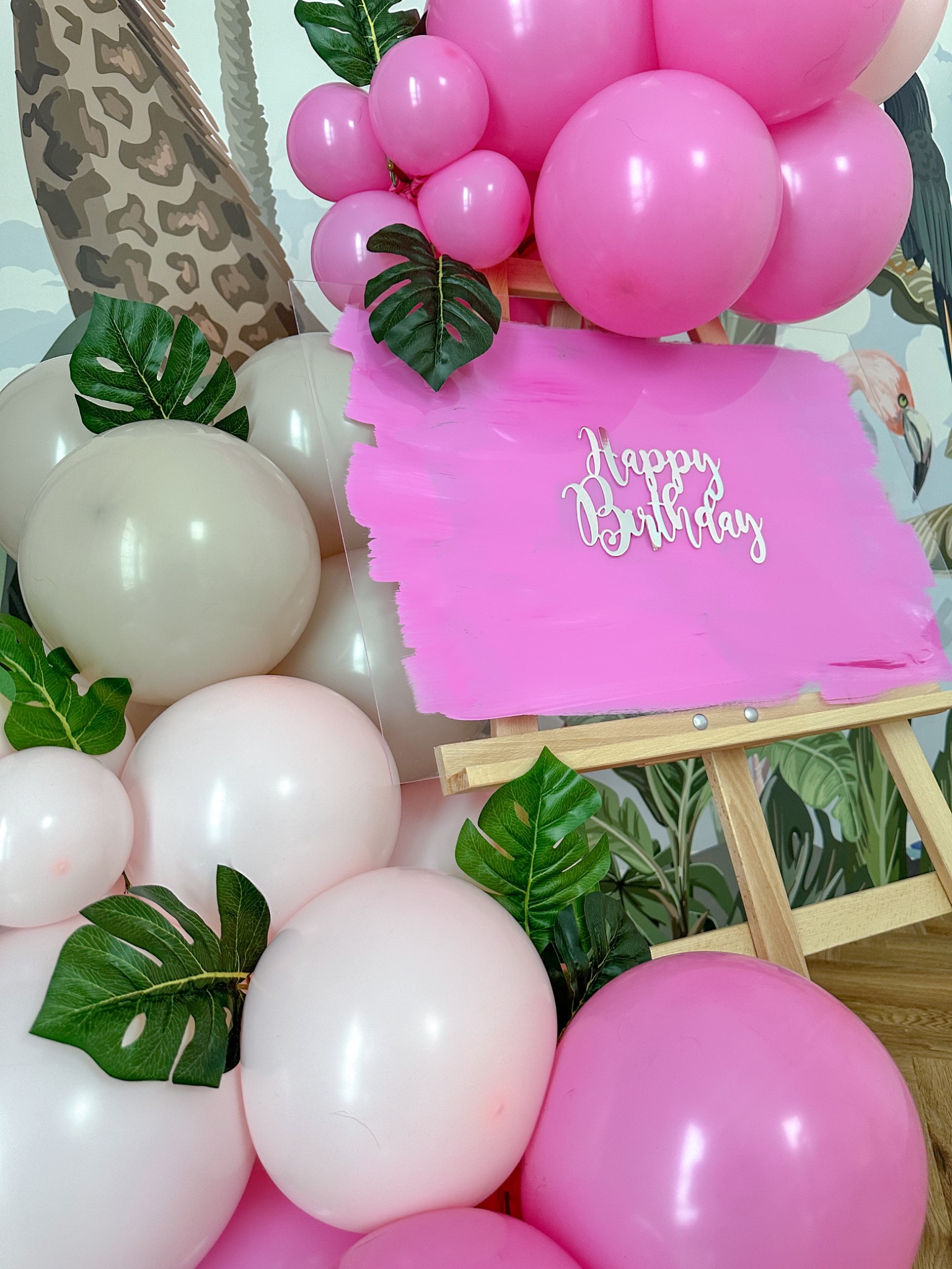 Dekoracja urodzinowa z różowych i białych balonów, zielonych liści i różowej tablicy z napisem 'Happy Birthday' na drewnianym sztaludze, na tle tapety w tropikalne wzory.