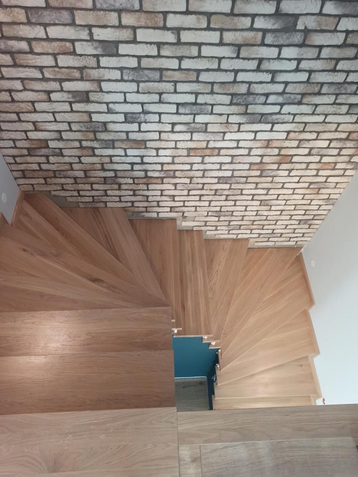 Kątowy widok z góry na drewniane schody spiralne z dębowymi stopniami, kontrastujące z ceglaną ścianą i białym fragmentem ściany, z widocznym fragmentem turkusowego pomieszczenia na dole.