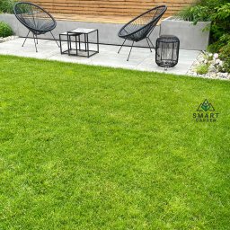 Your Smart Garden Sp. z o.o. - Nowoczesny ogród z idealnie przystrzyżonym trawnikiem, dwoma czarnymi fotelami typu Acapulco, stolikiem kawowym z marmurowym blatem i drewnianym panelem ogrodzeniowym w tle.