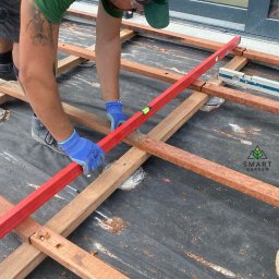 Your Smart Garden Sp. z o.o. - Pracownik w zielonej koszulce i słuchawkach poziomuje czerwoną poziomicą drewniany szkielet tarasu ułożony na czarnej geowłókninie.