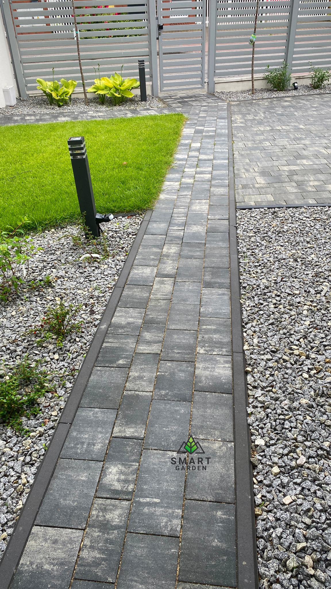 Szary chodnik z kostki brukowej obramowany ciemniejszymi obrzeżami, otoczony drobnym żwirem i trawnikiem, prowadzący do szarej bramy w tle.