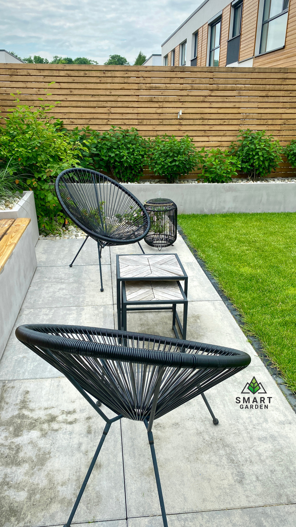 Nowoczesny ogród z drewnianym ogrodzeniem, betonowym podwyższeniem z nasadzeniami, trawnikiem oraz zestawem mebli ogrodowych: dwoma czarnymi krzesłami z plecionki i stolikiem kawowym z drewnianym...