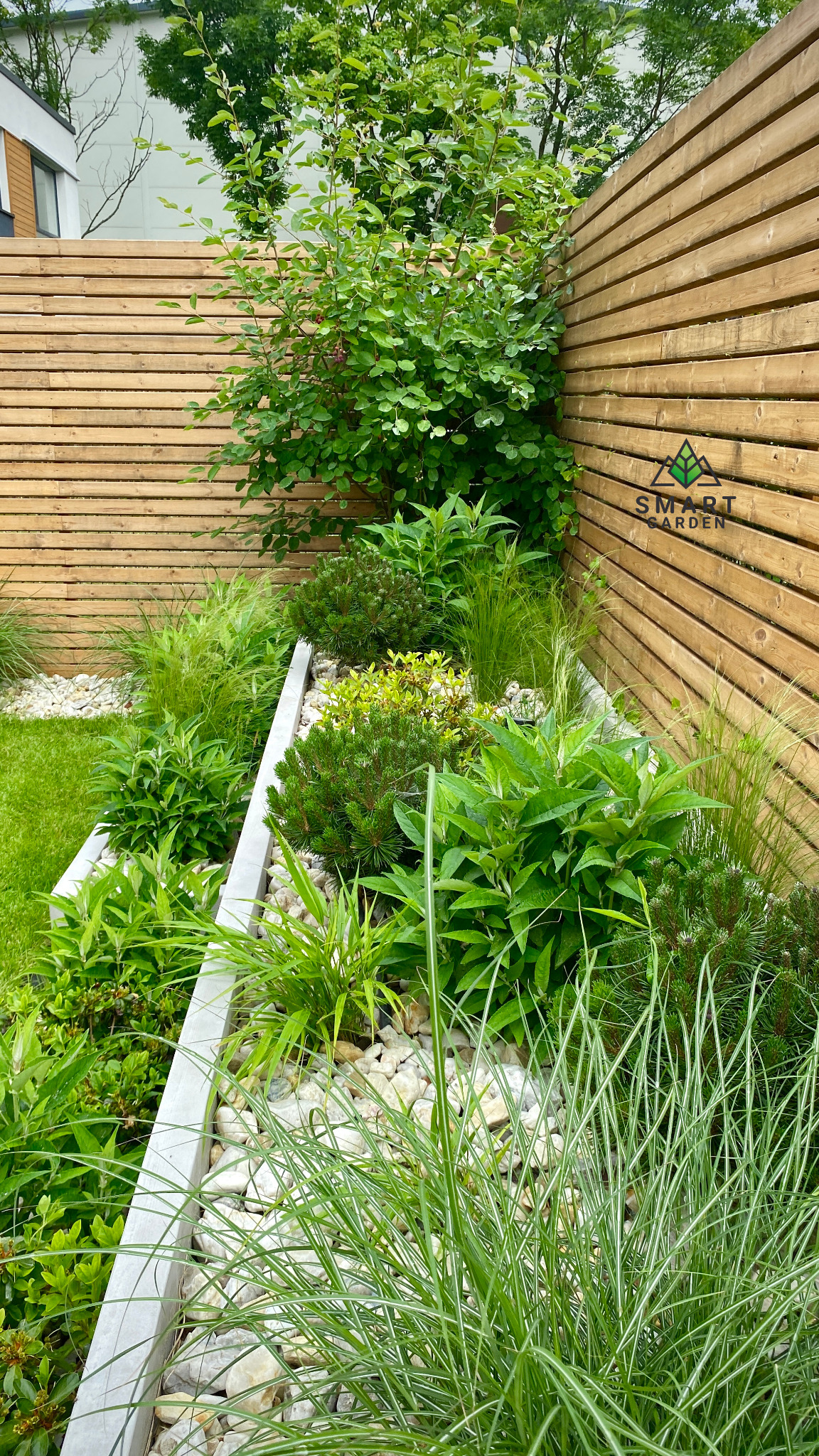 Nowoczesny ogród z drewnianym panelem, różnorodne rośliny w korycie wysypanym białym kamieniem, widoczne logo firmy Smart Garden na panelu.