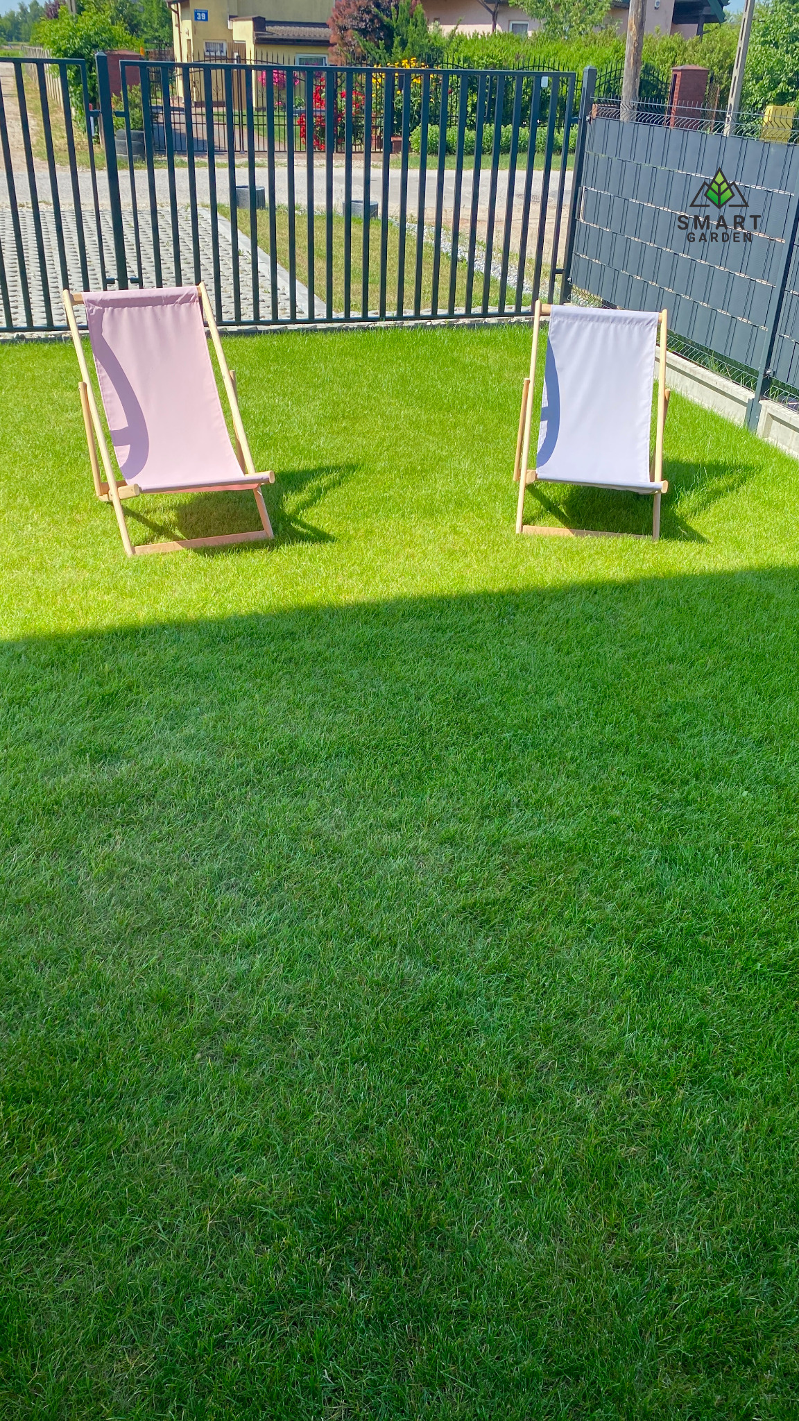 Równy trawnik z dwoma leżakami w odcieniach różu i szarości, ogrodzony czarnym metalowym płotem i szarą osłoną z logo Smart Garden.