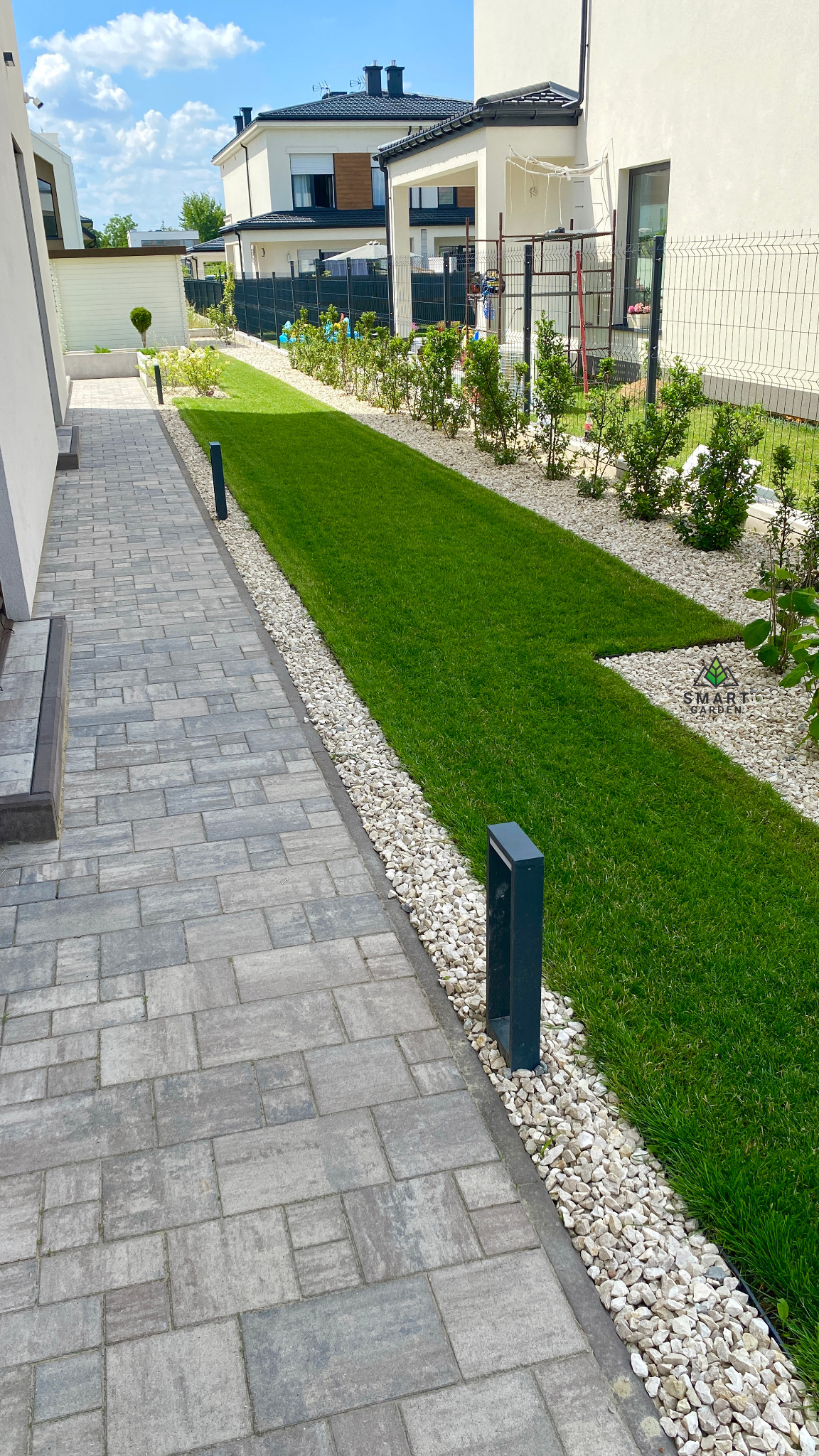 Nowoczesny ogród przydomowy z równo przystrzyżonym trawnikiem, obrzeżami z białych kamieni, szarą kostką brukową na ścieżce i niskimi lampami oświetlającymi, w tle zabudowa jednorodzinna.