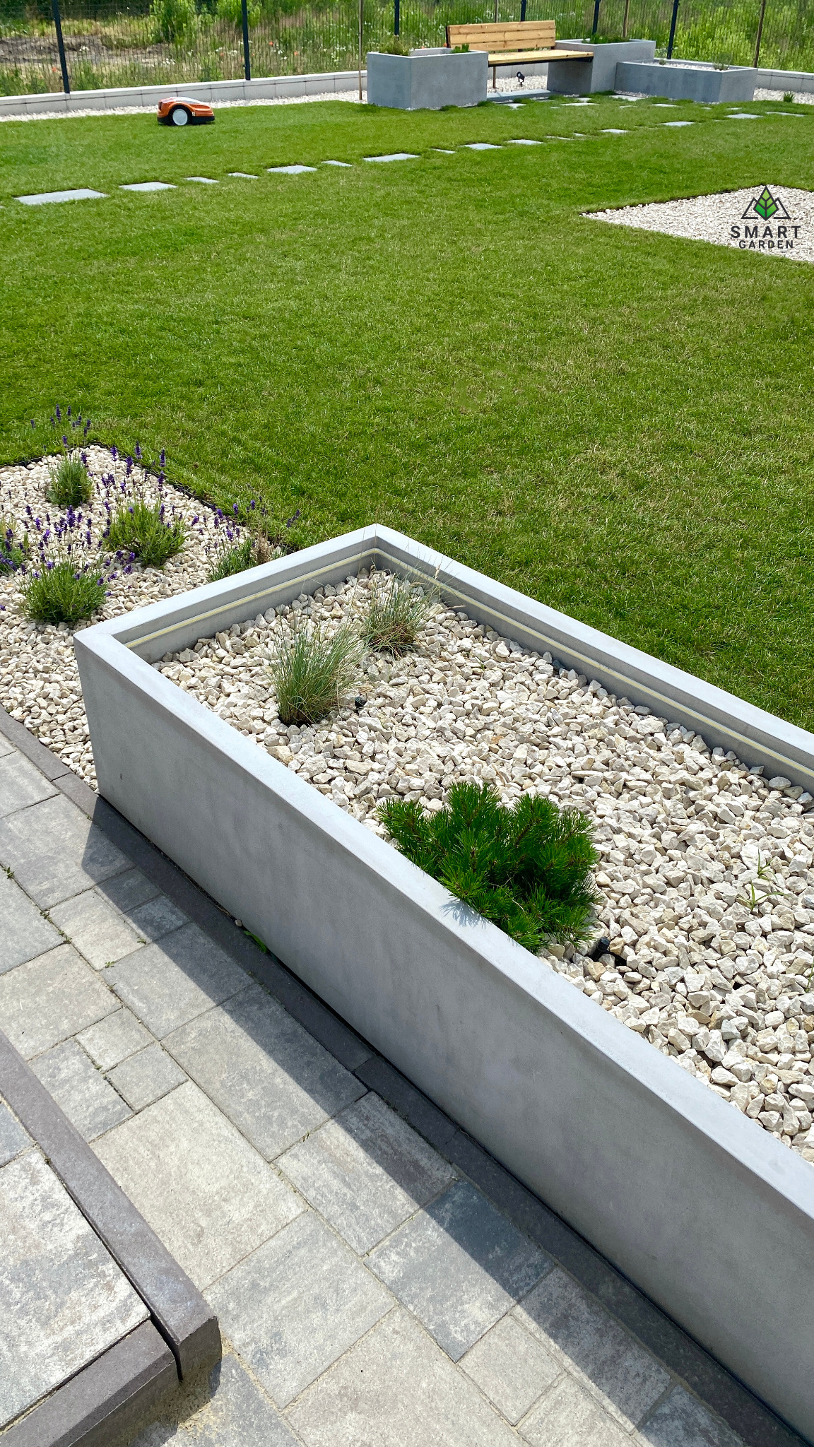 Nowoczesny ogród z betonowymi rabatami wypełnionymi białym żwirem i niskimi roślinami, na trawniku robot koszący.