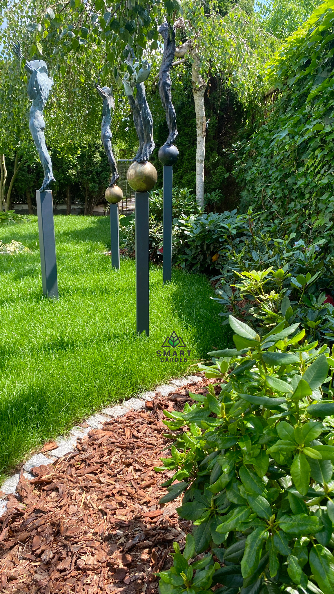 Ogród z rzeźbami figuralnymi na metalowych postumentach, trawnik, kora na rabatach i różnorodne rośliny ozdobne.