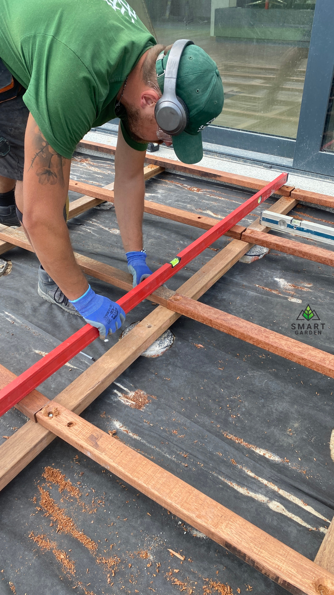 Pracownik w zielonej koszulce i słuchawkach poziomuje czerwoną poziomicą drewniany szkielet tarasu ułożony na czarnej geowłókninie.