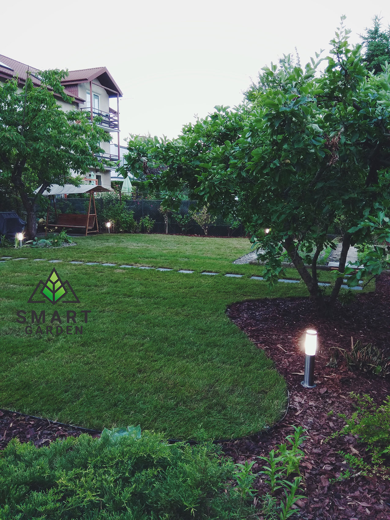 Zadbany ogród z równo przystrzyżonym trawnikiem, drzewami owocowymi, podświetlonymi ścieżkami i elementami dekoracyjnymi z kory drzewnej, oświetlenie ogrodowe w postaci lamp stojących.